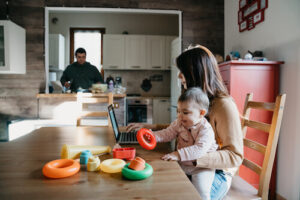 A woman holds an infant in her lap while she uses a laptop at the dining table