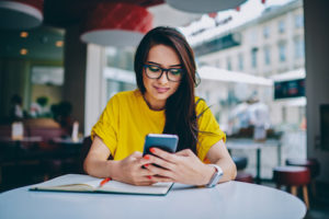 A woman in a yellow shirt sits at a desk and looks at her cell phone