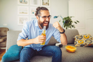 Man cheering on living room couch drinking beer and snacking on chips while watching The Draft