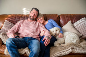 Husband, wife, and dog cuddling together on couch while watching movie on 4K TV