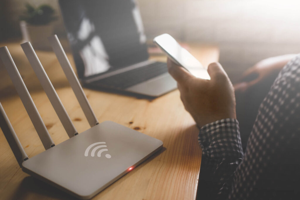 A router and a laptop sit on a desk. A hand is hold a cellphone