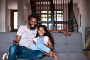 Dad and daughter sitting and smiling on living room couch watching TV