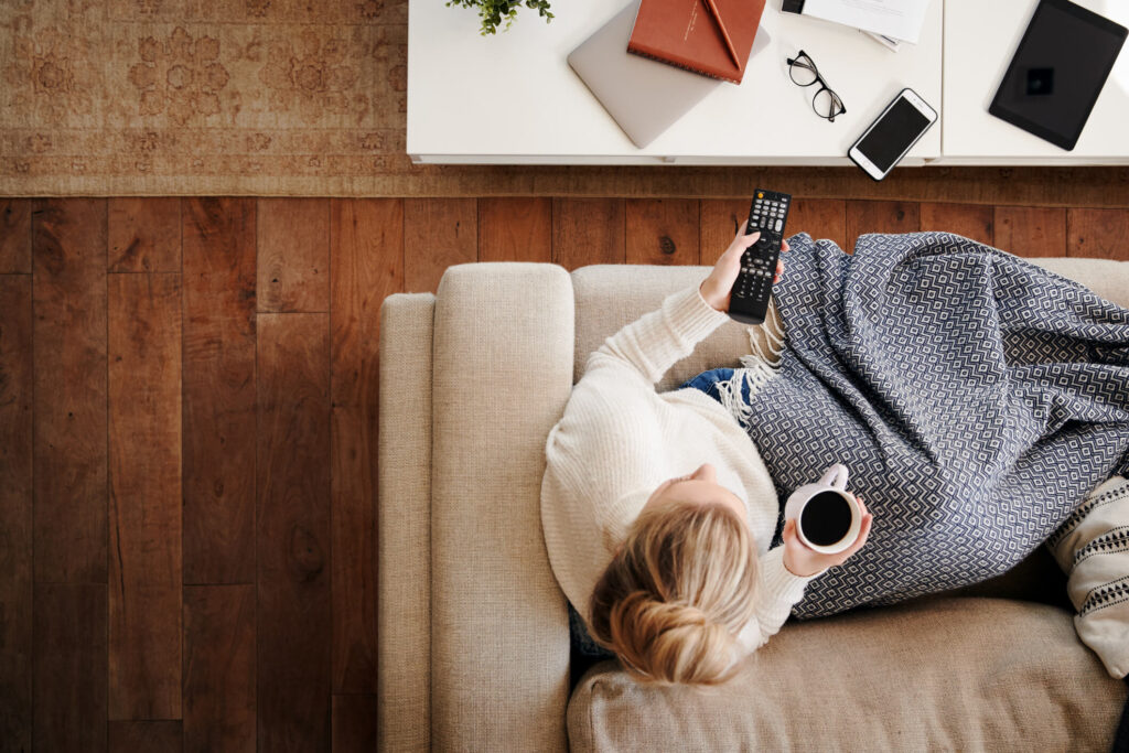 Overhead Shot Looking Down On Woman At Home Lying On Sofa, Drinking Coffe, and Watching TV