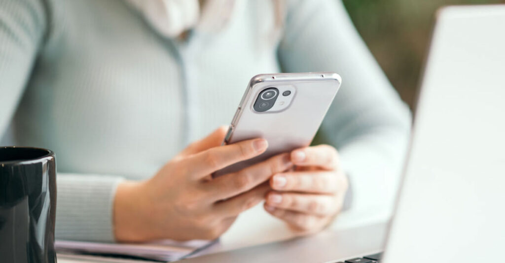 Woman holds cellphone while working on a laptop