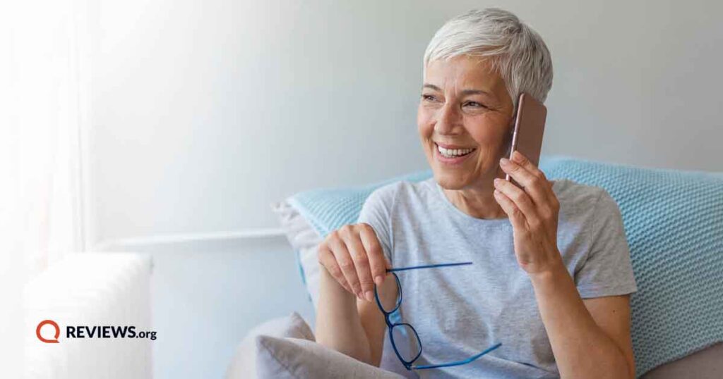 Happy senior woman holding glasses talking on phone while sitting near laptop