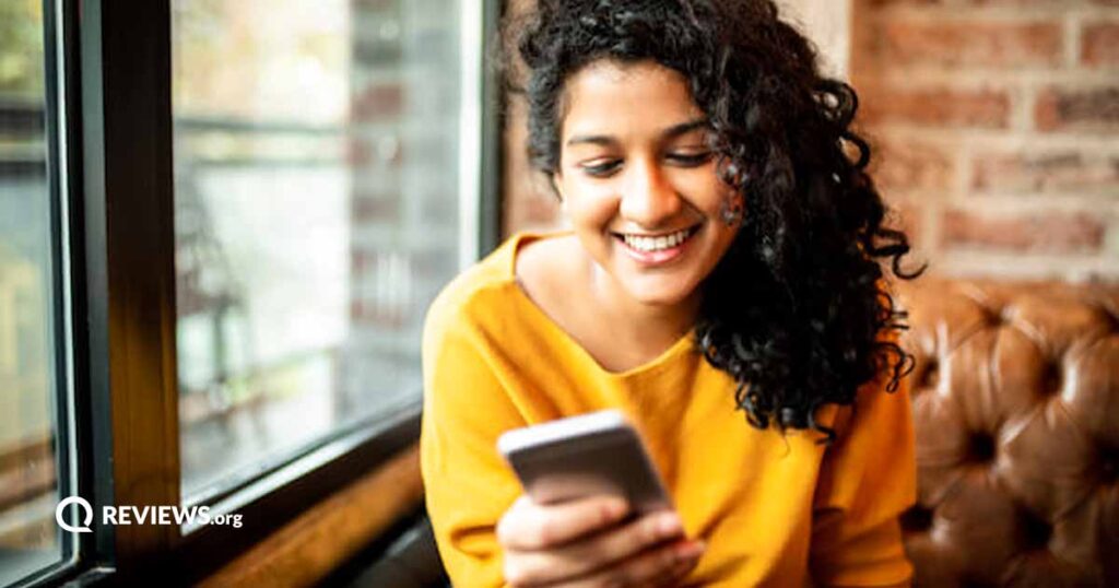 A mixed race woman with long curly hair and a yellow shirt surfs the web on her cellphone