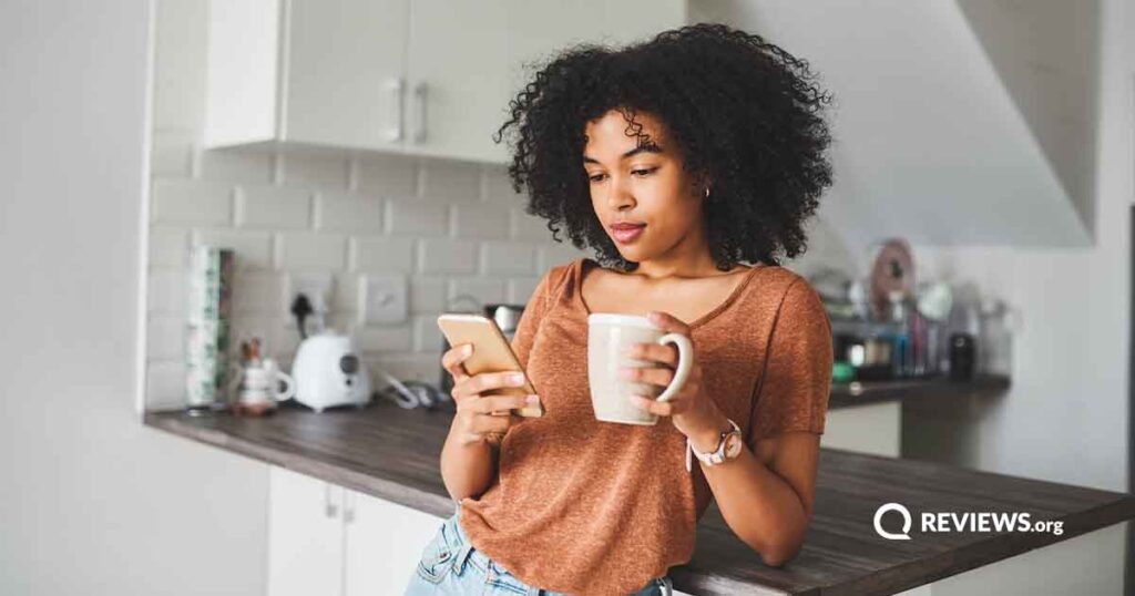 A woman looks at her phone while drinking coffee in the kitchen.
